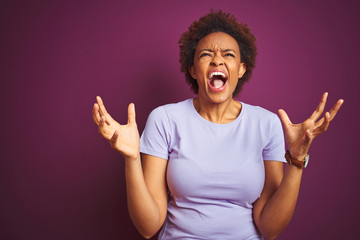 Young beautiful african american woman with afro hair over isolated purple background crazy and mad shouting and yelling with aggressive expression and arms raised. Frustration concept.