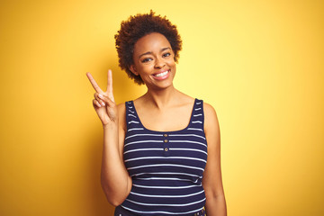Beauitul african american woman wearing summer t-shirt over isolated yellow background smiling looking to the camera showing fingers doing victory sign. Number two.