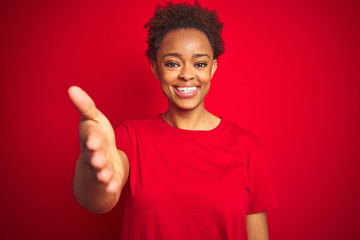 Young beautiful african american woman with afro hair over isolated red background smiling friendly offering handshake as greeting and welcoming. Successful business.