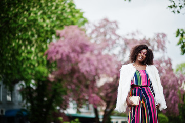 Fototapeta premium Fashionable african american woman in pink striped jumpsuit with fluffy faux fur coat posed at spring bloom street.