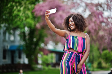 Fashionable african american woman in pink striped jumpsuit posed at spring bloom street and making selfie by mobile phone.