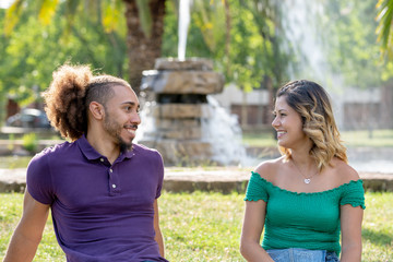 Obraz premium happy young couple in love looking at each other smiling sitting in front of a fountain on the park lawn