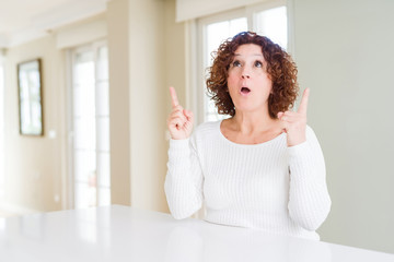 Beautiful senior woman wearing white sweater at home amazed and surprised looking up and pointing with fingers and raised arms.