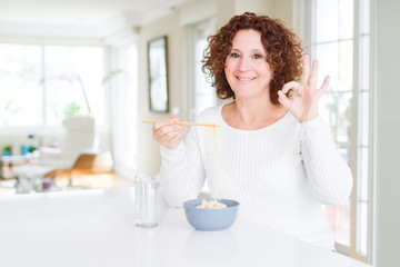 Senior woman eating asian noodles using chopsticks doing ok sign with fingers, excellent symbol