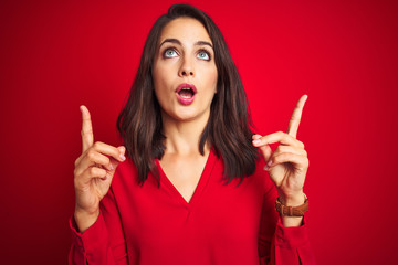 Young beautiful woman wearing shirt standing over red isolated background amazed and surprised looking up and pointing with fingers and raised arms.