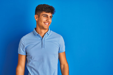 Fototapeta premium Young indian man wearing casual polo standing over isolated blue background looking away to side with smile on face, natural expression. Laughing confident.