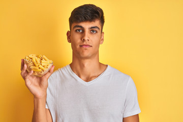 Young indian man holding bowl with dry pasta standing over isolated yellow background with a confident expression on smart face thinking serious