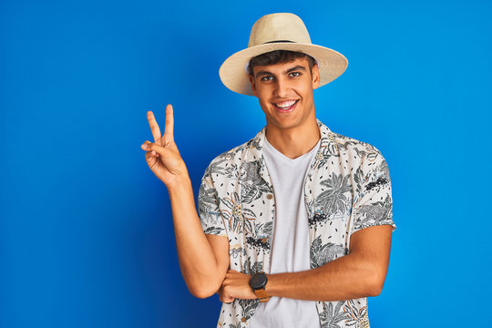 Indian man on vacation wearing hawiaian shirt summer hat over isolated blue background smiling with happy face winking at the camera doing victory sign. Number two.
