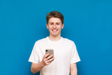 A lucky boy on a blue background holding a smartphone in his hands and smiling while looking at the camera. A lucky young man uses a smartphone stands on a blue background.