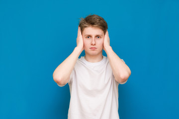 A mysterious man in a white T-shirt covers his ears with his hands, against a blue background. Blue background.