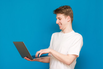 Portrait of a cheerful man, standing on a blue background, holding a laptop in his hands, playing a game. A man with a laptop, wearing a white T-shirt, is isolated on a blue background. - obrazy, fototapety, plakaty