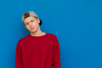 A handsome guy in stylish red clothes stands against a blue background, smiles and looks at the camera, wears a cap. Portrait of a joyful young man in blue background