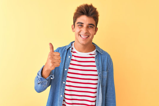 Young handsome man wearing striped t-shirt and denim shirt over isolated yellow background doing happy thumbs up gesture with hand. Approving expression looking at the camera showing success.