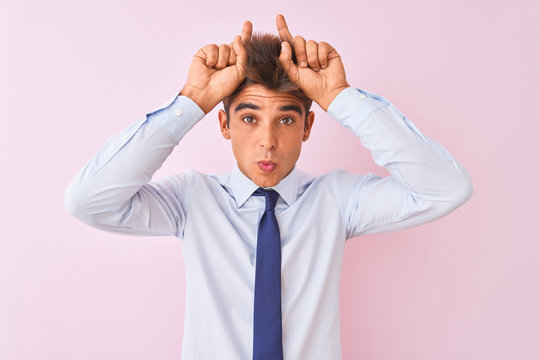 Young handsome businessman wearing shirt and tie standing over isolated pink background doing funny gesture with finger over head as bull horns