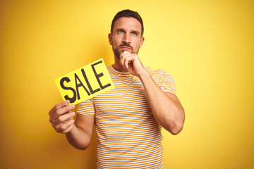 Young man holding sale advertising poster board over yellow isolated background serious face...