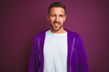 Young fitness man wearing casual sports sweatshirt over purple isolated background winking looking at the camera with sexy expression, cheerful and happy face.