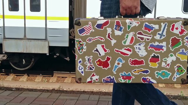the passenger is on the platform of the station with a suitcase with stickers from different countries in his hand