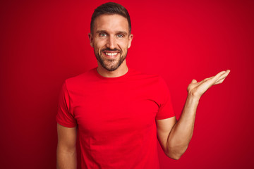 Young handsome man wearing casual t-shirt over red isolated background smiling cheerful presenting and pointing with palm of hand looking at the camera.