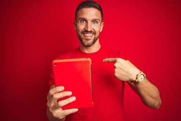 Young man using digital touchpad tablet over red isolated background with surprise face pointing finger to himself