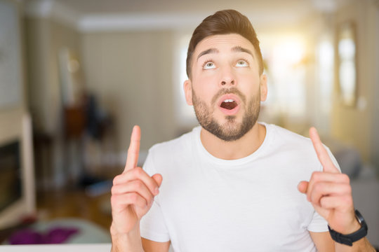 Young handsome man wearing casual white t-shirt at home amazed and surprised looking up and pointing with fingers and raised arms.