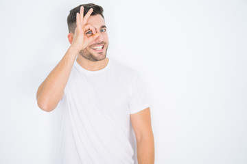 Young handsome man wearing casual white t-shirt over white isolated background doing ok gesture with hand smiling, eye looking through fingers with happy face.