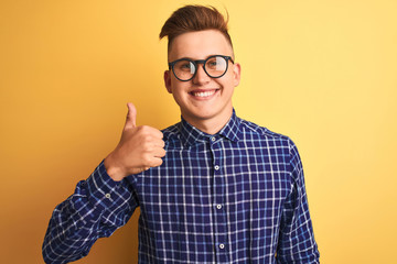 Young handsome man wearing casual shirt and glasses over isolated yellow background doing happy thumbs up gesture with hand. Approving expression looking at the camera with showing success.