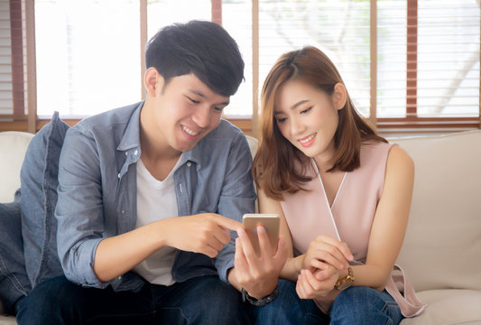 Young Asian Couple Or Friend Looking Smart Mobile Phone With Enjoy At Living Room, Happy Family Relax And Fun Media Entertainment On Smartphone On Sofa Together At Home, Lifestyle Concept.