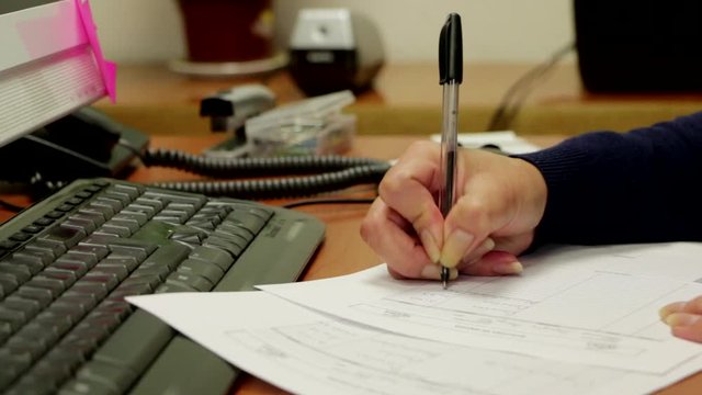 Detalle de una mano de mujer escribiendo con un bol&iacute;grafo en documentos de oficina frente a una computadora