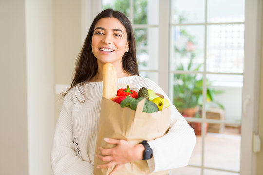 Beautiful young woman smiling holding a paper bag full of fresh groceries at home