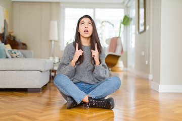 Young beautiful woman sitting on the floor at home amazed and surprised looking up and pointing with fingers and raised arms.