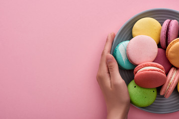 cropped view of woman holding plate with multicolored delicious French macaroons on pink background with copy space