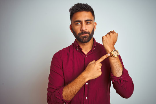 Young indian man wearing red elegant shirt standing over isolated grey background In hurry pointing to watch time, impatience, looking at the camera with relaxed expression