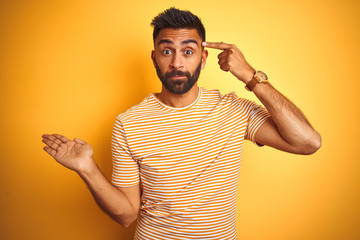 Young indian man wearing t-shirt standing over isolated yellow background confused and annoyed with open palm showing copy space and pointing finger to forehead. Think about it.