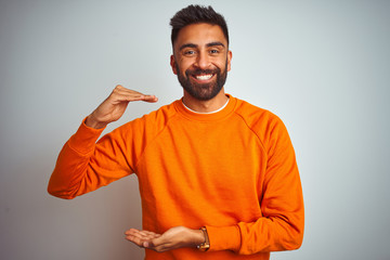 Young indian man wearing orange sweater over isolated white background gesturing with hands showing big and large size sign, measure symbol. Smiling looking at the camera. Measuring concept.