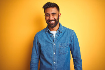 Young indian man wearing denim shirt standing over isolated yellow background winking looking at the camera with sexy expression, cheerful and happy face.