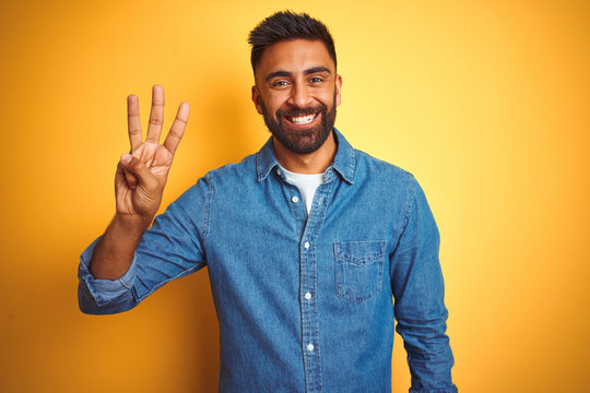 Young indian man wearing denim shirt standing over isolated yellow background showing and pointing up with fingers number three while smiling confident and happy.