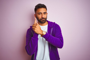 Young indian man wearing purple sweatshirt standing over isolated pink background Holding symbolic gun with hand gesture, playing killing shooting weapons, angry face
