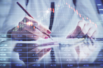 A woman hands writing information about stock market in notepad. Forex chart holograms in front. Concept of research.