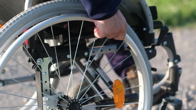 An old man with a disability turns the gray wheel of a wheelchair with his hands and slowly goes forward on a dirt road. Close up.