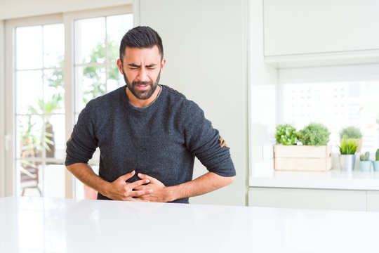 Handsome hispanic man wearing casual sweater at home with hand on stomach because indigestion, painful illness feeling unwell. Ache concept.