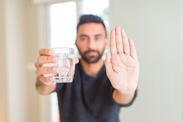 Handsome hispanic man drinking a fresh glass of water with open hand doing stop sign with serious and confident expression, defense gesture