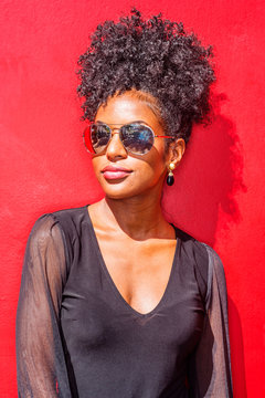 Portrait Of Young African American Woman In New York City. Young Black Woman With Afro Hairstyle Wearing Long Sleeve Mesh Sheer Shirt, Sunglasses, Standing Against Red Background Under Sun..