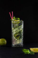 Fresh summer cucumber lemonade with two pink straws in the glass. Black background. Lime and mint on the table.