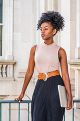 Obraz na płótnie Canvas Young beautiful African American woman with afro hairstyle wearing sleeveless light color top, belt, black skirt, holding laptop computer, standing in vintage office building in New York, thinking..