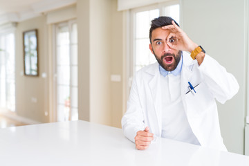 Handsome hispanic doctor or therapist man wearing medical coat at the clinic doing ok gesture shocked with surprised face, eye looking through fingers. Unbelieving expression.