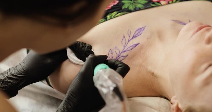Creating tattooing in tattoo salon. Tattoo artist makes tattoo with leaves in studio for young woman, close up. She uses professional tattoo machine making her job. Painting body art image on skin.