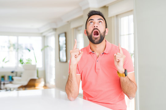 Handsome hispanic man wearing casual t-shirt at home amazed and surprised looking up and pointing with fingers and raised arms.