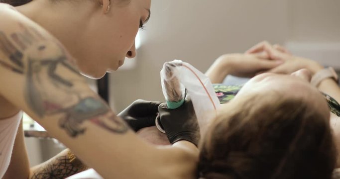 Tattoo artist making body art picture in studio. Profesional master young girl creating tattoo with leaves for woman on shoulder, closeup. Tattooist making tattooing machine nature pattern.