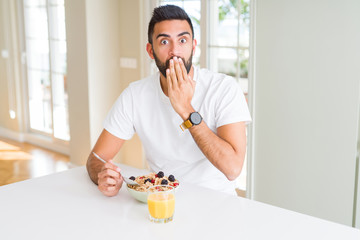 Handsome hispanic man eating healthy breakfast in the morning at home cover mouth with hand shocked with shame for mistake, expression of fear, scared in silence, secret concept