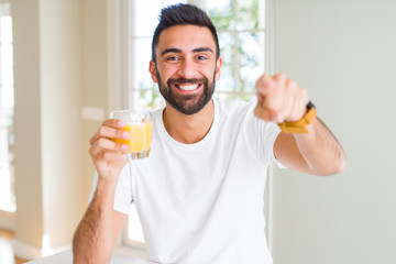 Handsome hispanic man drinking healthy orange juice pointing with finger to the camera and to you,...